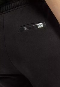 Czarne dresy wykonane z miękkiego materiału; mają małą kontrastową kieszonkę z błyszczącym czarnym wykończeniem oraz metką marki z tyłu.