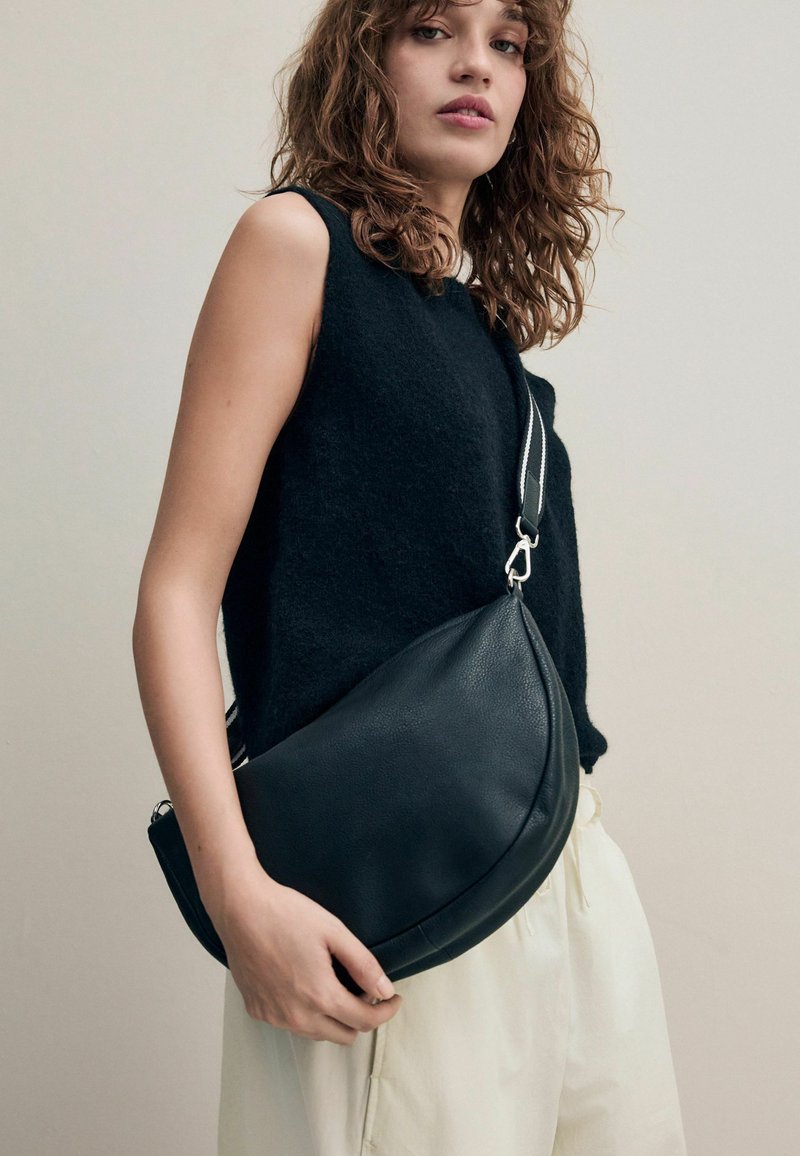 Sac à bandoulière en cuir noir avec une forme arrondie, doté d'un détail en métal argenté et d'une sangle réglable, tenu par une personne portant un haut sans manches.