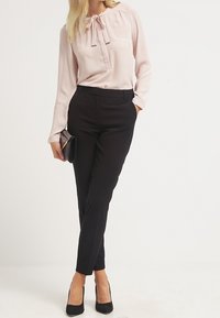 Žena v světlé růžové blůze s mašlí, černých elegantních kalhotách, černých lodičkách, držící černou psaníčko, stojící na bílé pozadí.
