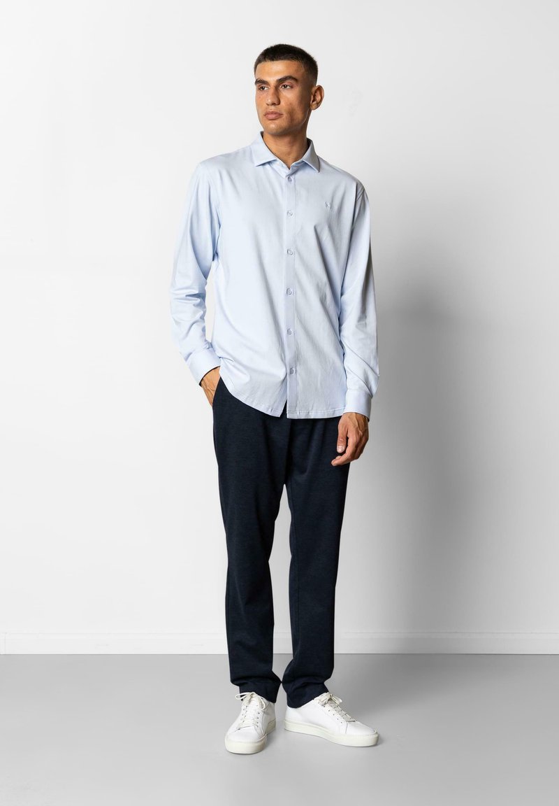 Chemise boutonnée bleu clair à manches longues, associée à un pantalon gris foncé et des baskets blanches, se tenant devant un arrière-plan neutre.