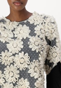 Grå blomstret topp med teksturerte kremfargede stoffblomster, prikkete mønster, rund halslinning og korte ermer.
