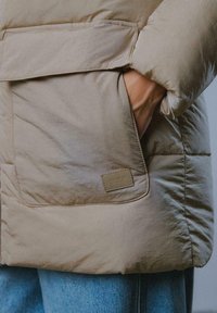 Beżowa puchowa kurtka z kieszenią z przodu, miękka w dotyku, z małą metką marki. Nosić ją na jasnoniebieskie dżinsy.