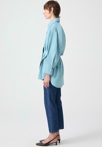 Camicia oversize azzurro chiaro con maniche lunghe, dettagli con bottoni e vita cinchata, abbinata a jeans dritti in denim scuro e scarpe slingback nere.