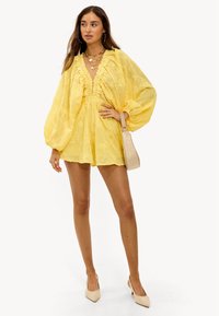 Combinaison jaune texturée avec manches bouffantes, encolure à volants et short. Associée à un petit sac et des escarpins à bout pointu de couleur neutre.