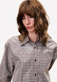 Vrouw met golvend bruin haar, gekleed in een bruin-wit geruit overhemd met pailletten, kijkt naar beneden tegen een effen achtergrond.