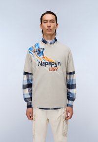 Lys grå t-shirt med en farverig skigrafik og teksten "Napapijri 1987", lagd over en blå og hvid ternet skjorte, iført med beige bukser.
