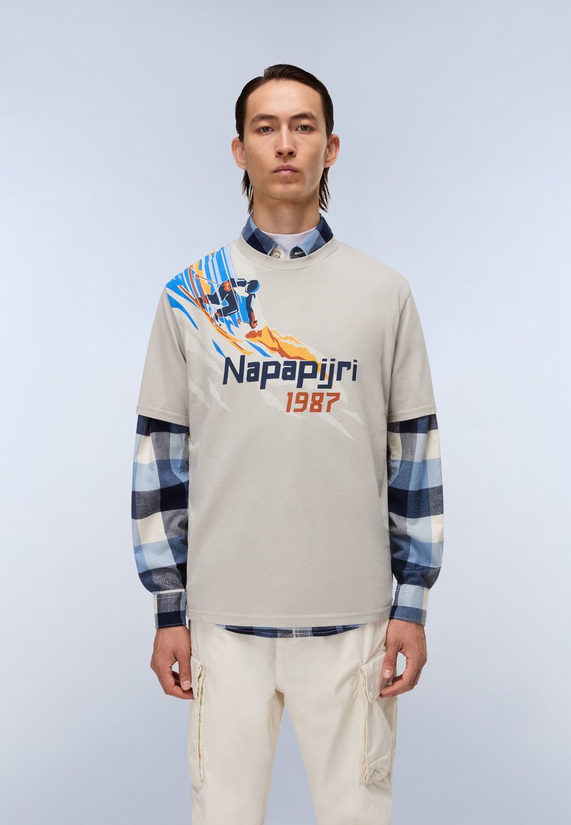 Lys grå t-shirt med en farverig skigrafik og teksten "Napapijri 1987", lagd over en blå og hvid ternet skjorte, iført med beige bukser.