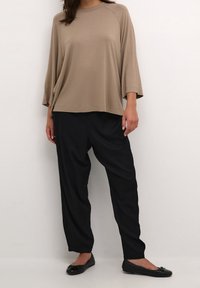 Žena v volném béžovém dlouhém rukávu, černých volně střižených kalhotách a černých balerínách stojící na bílém pozadí.