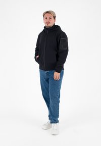 Giacca nera con cappuccio, apertura con zip, polsini elasticizzati e tasche laterali; indossata sopra un dolcevita nero, abbinata a jeans blu e scarpe da ginnastica bianche.