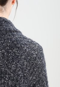 Teksturowany ciemnoszary kardigan z luźnym splotem, w którym znajdują się jaśniejsze szare nitki, co nadaje mu przytulny i miękki wygląd.