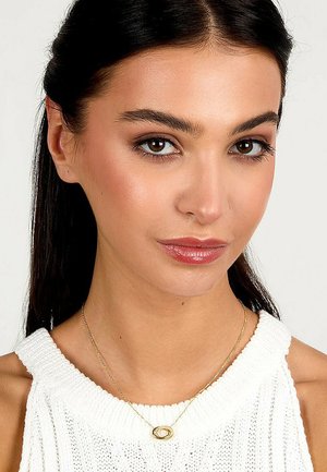 Goldene Halskette mit einem ovalen Anhänger, zarter Kette und glatter Textur, getragen über einem weißen Strickoberteil. Der Hautton ist warm.