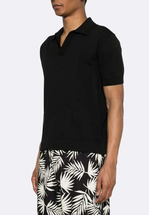 Uomo che indossa una camicia nera a maniche corte con colletto e pantaloncini neri con motivi di foglie tropicali bianche, in piedi di profilo.