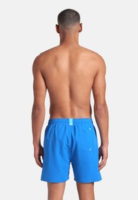 Uomo con capelli corti in piedi di spalle, indossando pantaloncini da bagno blu brillante con vita elastica e una tasca sul retro, su sfondo bianco.