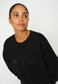 Čierna mikina s okrúhlym výstrihom. Na prednej strane sa nachádza kamienkový dizajn krídel a symbolov. Mäkka tkanina, voľný strih.