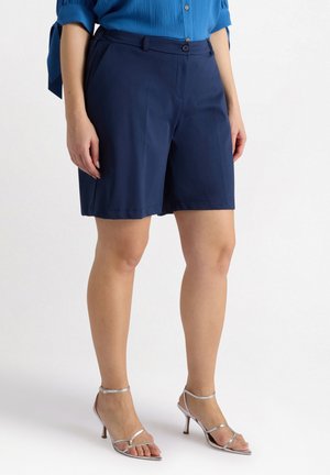 Marineblauwe shorts met een vlakke voorkant, knoopsluiting en riemlussen. Glad stof, kniehoogte, gecombineerd met zilveren sandaaltjes met bandjes.