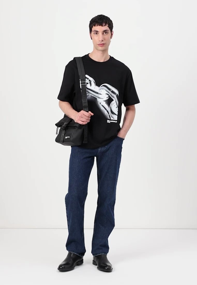 T-shirt noir avec un motif graphique, associé à un jean bleu et des bottines noires. Un petit sac bandoulière noir est porté sur l'épaule.