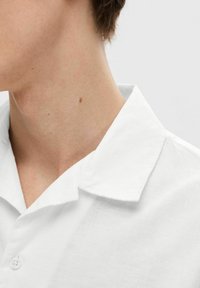 Balta lininė marška su atlapų apykakle ir sagčių uždarymu, demonstruojanti lengvą ir šiek tiek permatomą tekstūrą.