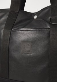 Sac de voyage en cuir noir avec une surface texturée, doté d'un logo en patch rectangulaire et de poignées en tissu robustes. Accent de fermeture à pression argentée.