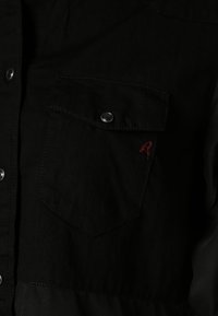 Camicia in denim nero con chiusura a bottoni, due tasche sul petto, bottoni a pressione argentati e un piccolo dettaglio ricamato rosso.