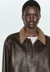 Veste en cuir marron avec un col en daim beige contrastant. Dotée d'une fermeture éclair à l'avant et d'une texture lisse. Le mannequin pose sur un fond uni.