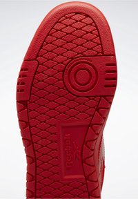 Czerwona podeszwa buta Reebok z diamentowym wzorem bieżnika, okrągłym designem uchwytu i tekstem "Podeszwa niebrudząca" w pobliżu podbicia.