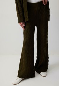 Alyvuogių žalios spalvos plačios kojų kelnės su kabeliu megztu tekstūru ir aukštu liemeniu, derinamos su baltomis raištinėmis bateliais.