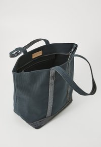 Tote bag en tissu bleu avec deux poignées, accents texturés gris et fermeture éclair. L'intérieur comprend une poche zippée et une étiquette de marque.