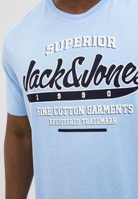 Světle modré bavlněné tričko s kulatým výstřihem. Obsahuje černobílý potisk textu: "SUPERIOR," "Jack & Jones," a "1990," s horizontální čarou.