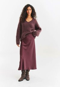 Žena s kudrnatými vlasy, oblečená v načechraném fialovém svetru, odpovídající dlouhé saténové sukni, hnědých botách a zlatém náhrdelníku, stojící na bílém pozadí.