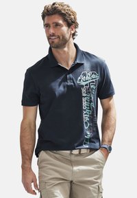 Marineblaues Polohemd mit kurzen Ärmeln, mit grafischem Text in Weiß und Türkis auf der Vorderseite. Kombiniert mit beigen Shorts und einem gemusterten Gürtel.