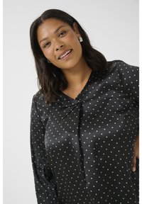 Blouse noire à pois avec une texture satinée, présentant un décolleté en V et un détail de nœud à l'avant. Le tissu a une finition lisse.