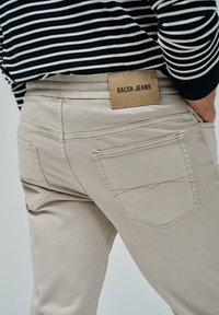 Pantalones de tejido elástico beige con cintura elástica y dos bolsillos traseros. Logo de "Salsa Jeans" en una etiqueta de cuero marrón claro.