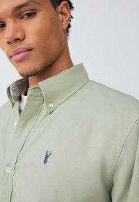 Camicia verde chiaro con bottoni e colletto, caratterizzata da un motivo ricamato blu navy sul petto e un dettaglio a righe all'interno del colletto.