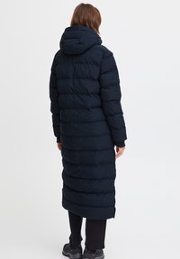 Lang, marineblå dunjakke med hette, quiltet design og elastiske cuffs. Har vertikale sømmer og en lett innsnevret silhuett.