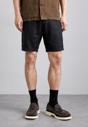 Sorte linned shorts med en afslappet pasform, parret med mørke sokker og brune ruskind loafers med frynse detaljer. Neutral baggrund.