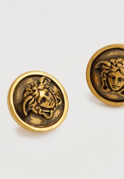Στρογγυλά σκουλαρίκια με χρυσό τόνο και ανάγλυφο σχέδιο κεφαλής Μέδουσας με σκούρο αντικέ φινίρισμα σε λείο μεταλλικό φόντο.