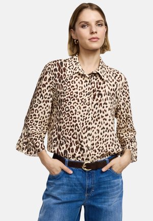 Blusa con stampa leopardata in tonalità beige e marroni, con scollo con colletto e maniche a campana. Abbinata a jeans blu e cintura marrone.