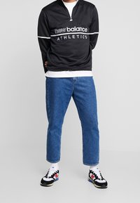 Černý sportovní top se čtvrtým zipem, bílým pruhovým akcentem a logem, doplněný modrými džínami a teniskami v černé, bílé a červené barvě.