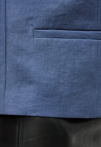 Κοντινό πλάνο σε τσέπη μπλε υφασμάτινου σακακιού πάνω από μαύρο υλικό δερματίνης με υφή από κάτω.