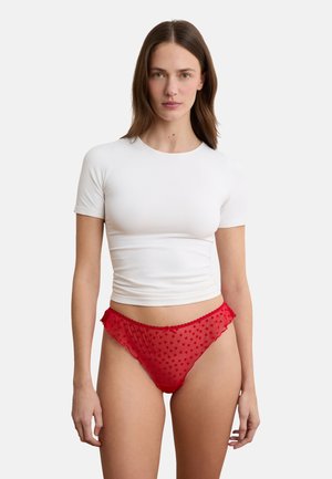 Femme portant une lingerie rouge transparente à pois et un crop top blanc ajusté à manches courtes, debout devant un fond blanc uni.