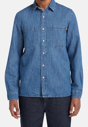 Muž nosící modrou riflovou košili s dlouhým rukávem, dvěma kapsami na hrudi a tmavými džínami, stojící s uvolněnými pažemi.