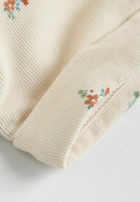 Krämfärgad ribbad tyg med blommönster i orange och turkos. Har en mjuk textur och design med vikta manschetter.