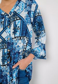 Blaue gemusterte Bluse mit V-Ausschnitt, Knopfverschluss und Glockenärmeln. Sie weist verschiedene florale und geometrische Designs in unterschiedlichen Blautönen auf.