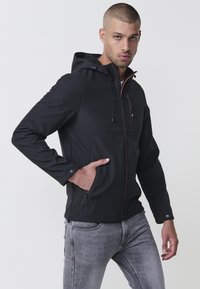 Jachetă neagră din material softshell, cu glugă, având un accent portocaliu la fermoar, buzunar pe piept cu fermoar, buzunare laterale și mansete ajustabile.