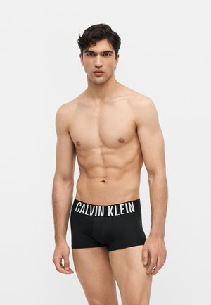Jeune homme en forme debout, portant un boxer Calvin Klein noir avec une ceinture blanche à logo, sur un fond clair uni.
