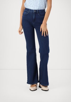 Frau trägt dunkelblaue hochtaillierte Schlagjeans, hellblaues Spitzentop und beige flache Schuhe und steht vor einem schlichten hellen Hintergrund.