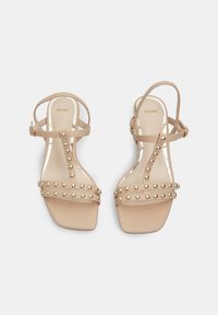 Krémové sandály na ploché podrážce s pásky, zdobené kovovými zlatými nýty podél pásků a s nastavitelnou sponou na kotníku.