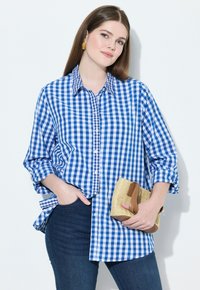 Camicia a quadri blu e bianca, maniche lunghe arrotolate, abbinata a jeans in denim scuro e una borsa a tracolla di paglia. Texture del tessuto liscia.