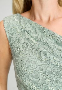 Robe en dentelle vert menthe avec un décolleté asymétrique, ornée de motifs floraux et de sequins réfléchissants, mettant en valeur des textures détaillées.