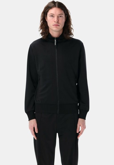 Giacca a track nera con zip, colletto alto, maniche lunghe e polsini a coste. Tessuto liscio, tasche laterali e design minimalista.